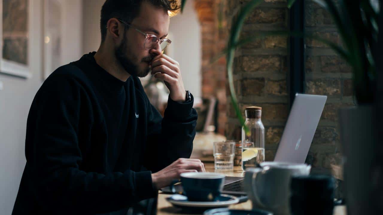 A man in glasses working on a laptop at a cafe.
