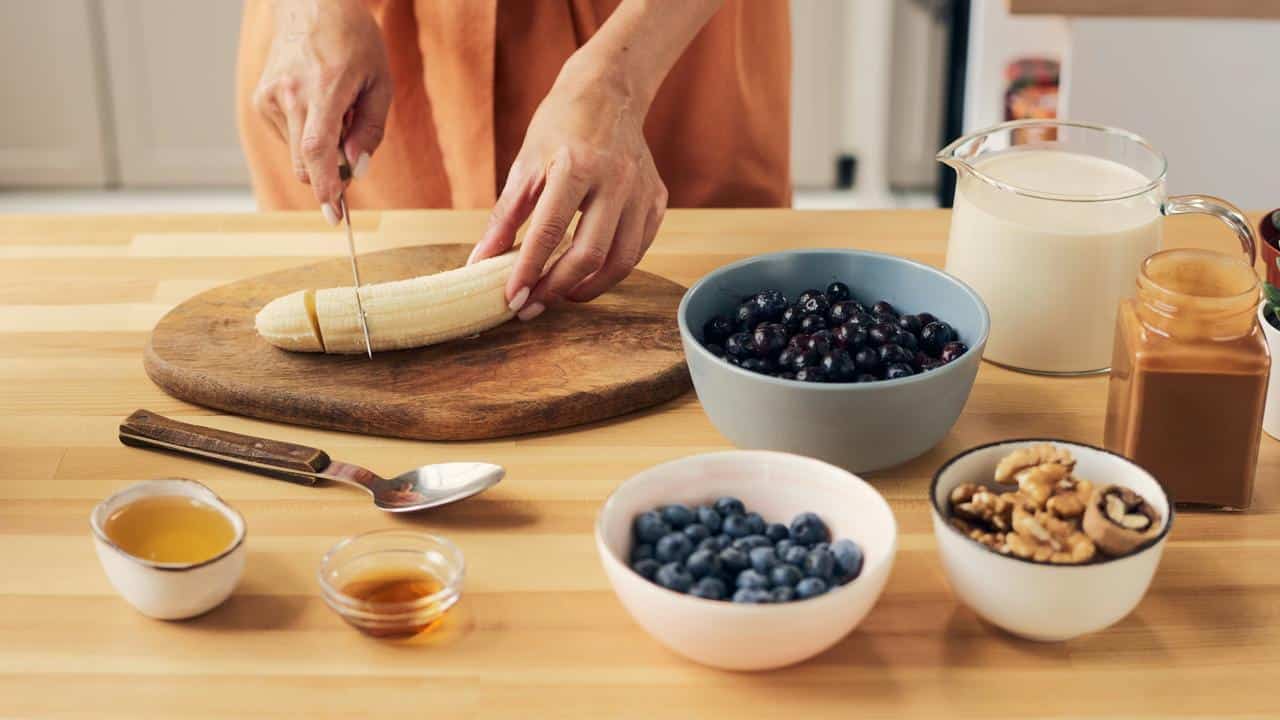 A person slicing a banana with fruits and nuts on the table.
