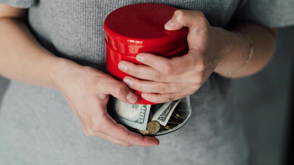 A person holding a red jar full of coins and cash.