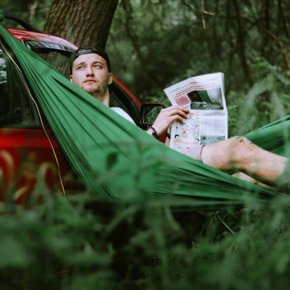 A man in a green hammock reading a newspaper in the forest.