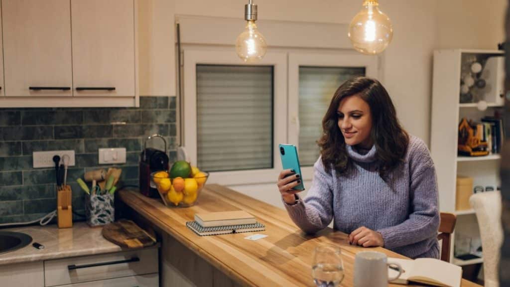 A happy woman is sitting in a kitchen and looking at her phone.