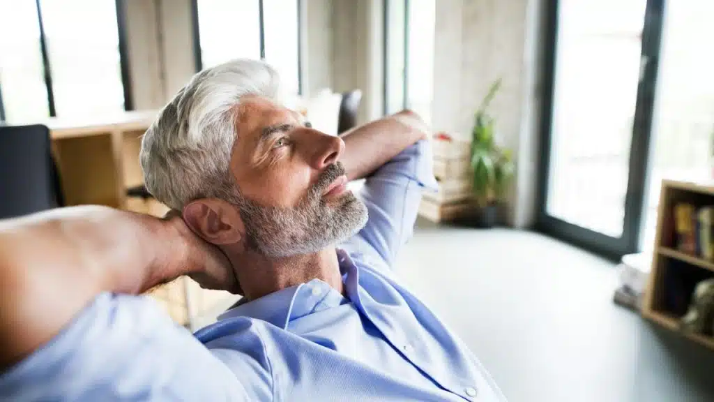 A thoughtful, gray-haired man in a light blue shirt leans back in a chair with his hands behind his head.