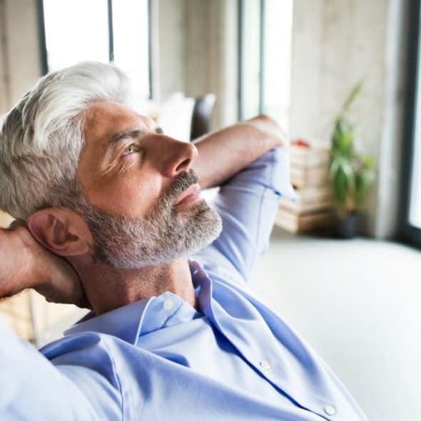 A thoughtful, gray-haired man in a light blue shirt leans back in a chair with his hands behind his head.