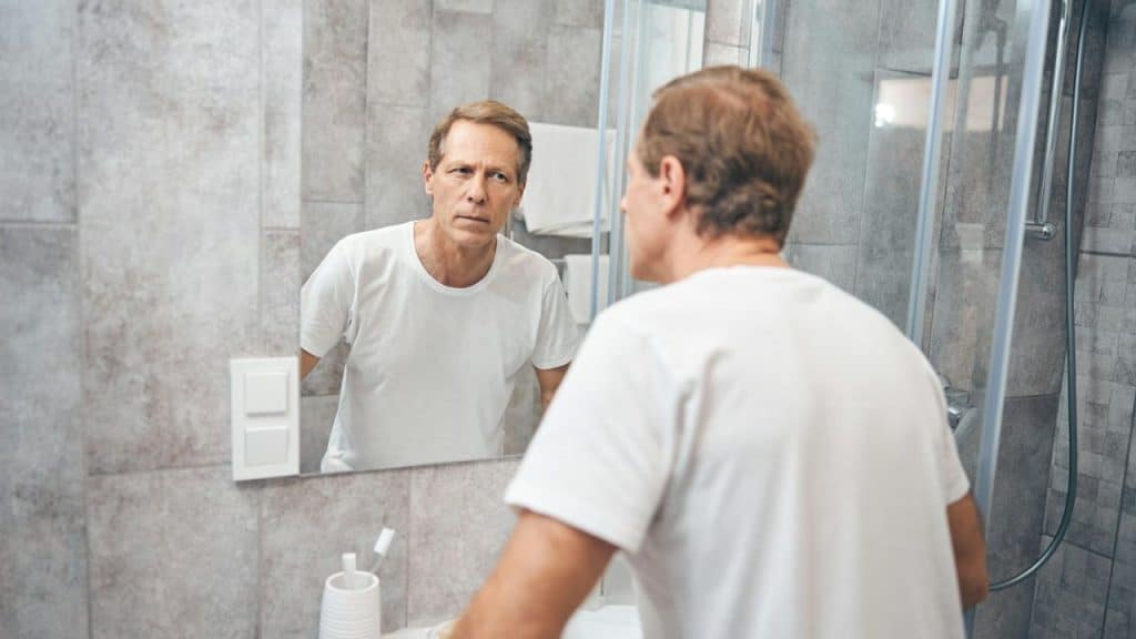 A concerned-looking middle-aged man stands in a bathroom, looking at his reflection in the mirror.