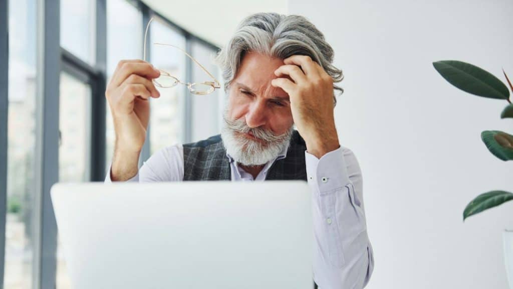A concerned-looking, gray-haired man with a beard sits at a desk, rubbing his forehead and holding glasses.