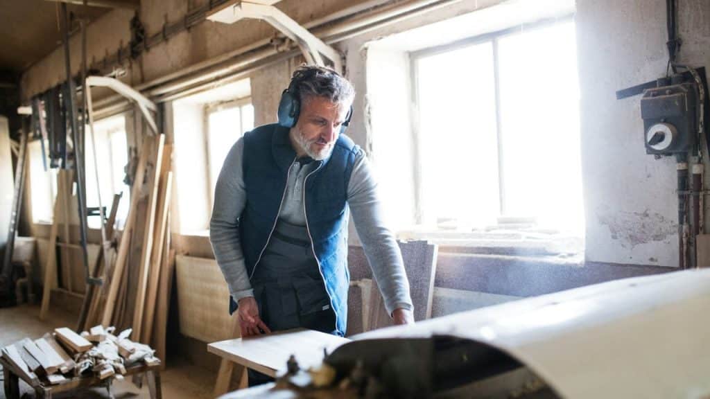 A focused, gray-haired man in earmuffs and a vest leans over a workbench in a bright workshop.
