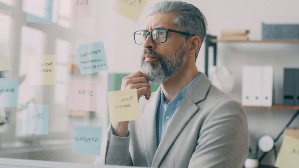 A handsome, gray-haired man in glasses and a blazer looks at a glass wall covered in sticky notes.