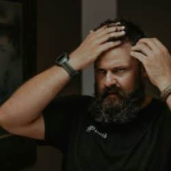 A worried man with a beard is looking in a mirror.