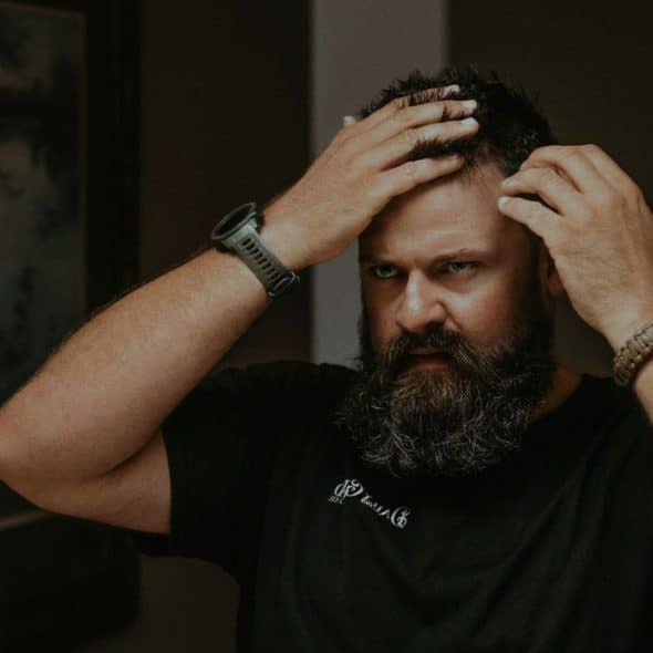 A worried man with a beard is looking in a mirror.