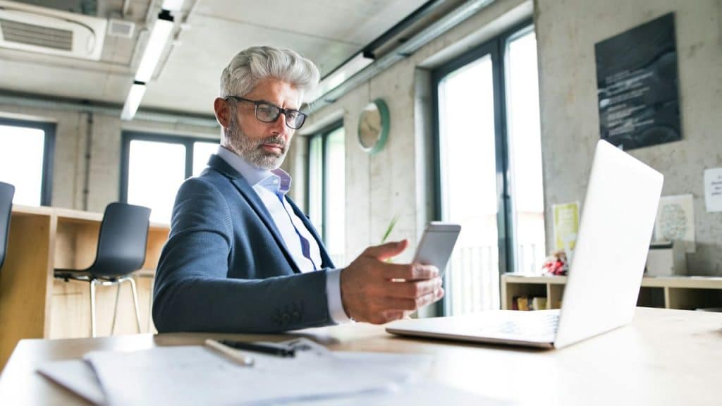 A mature, gray-haired man in a suit looks at his phone while working at his desk.