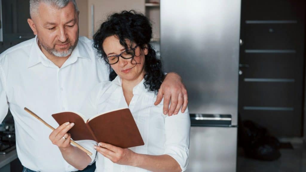 A mature couple stands together in a kitchen, looking at a notebook and holding each other.