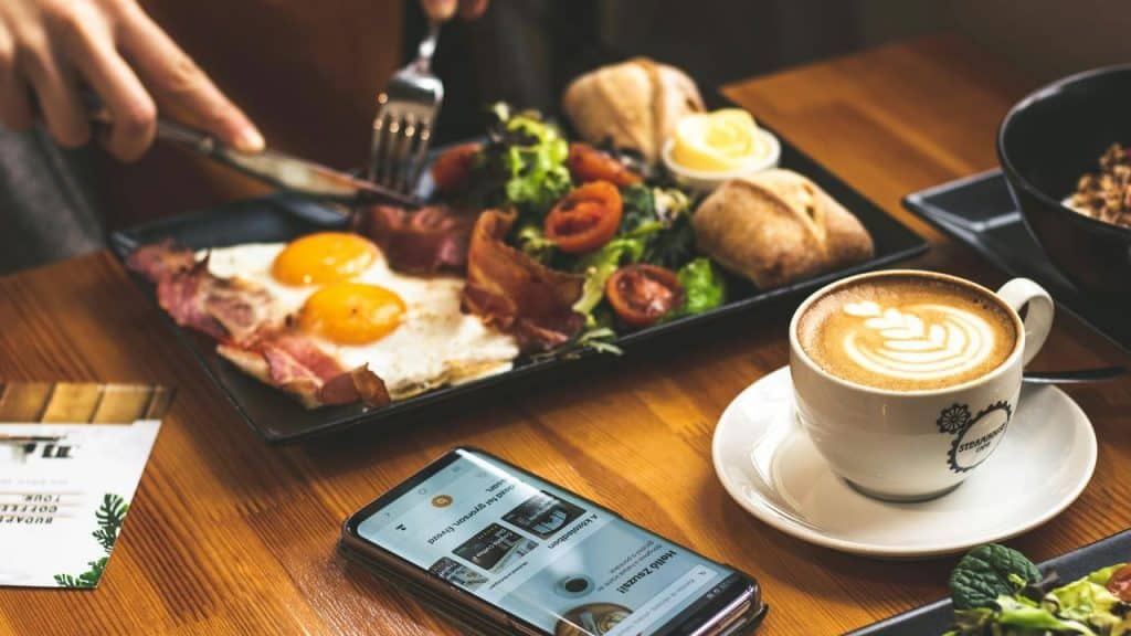 A breakfast plate with eggs, bacon, salad, bread, and a cup of latte.