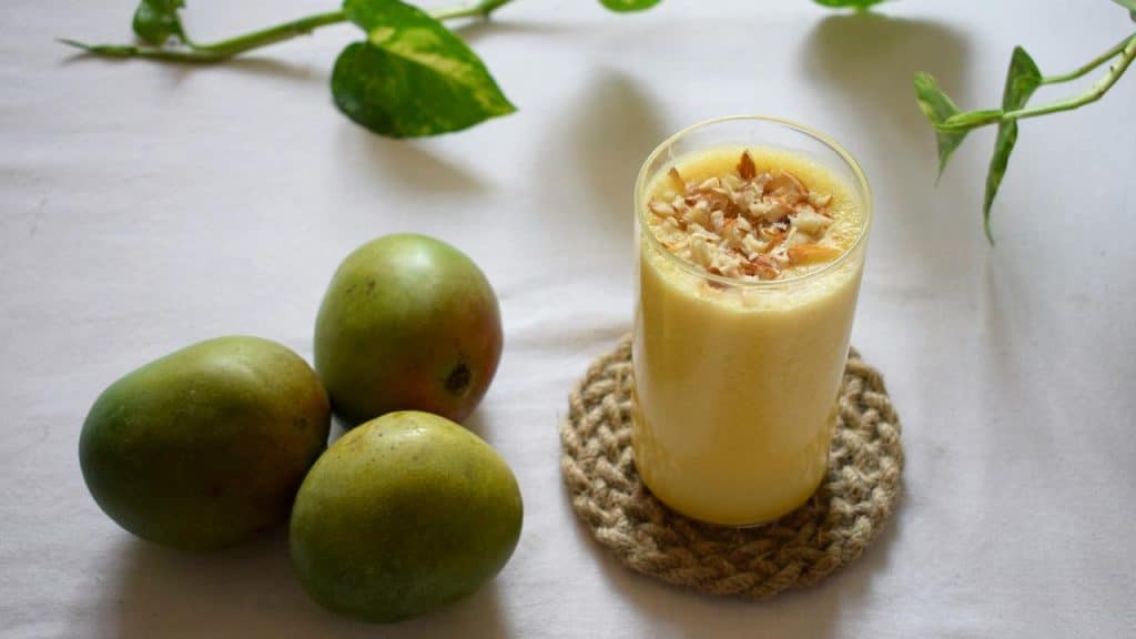 A glass of mango smoothie topped with nuts beside three whole mangoes.