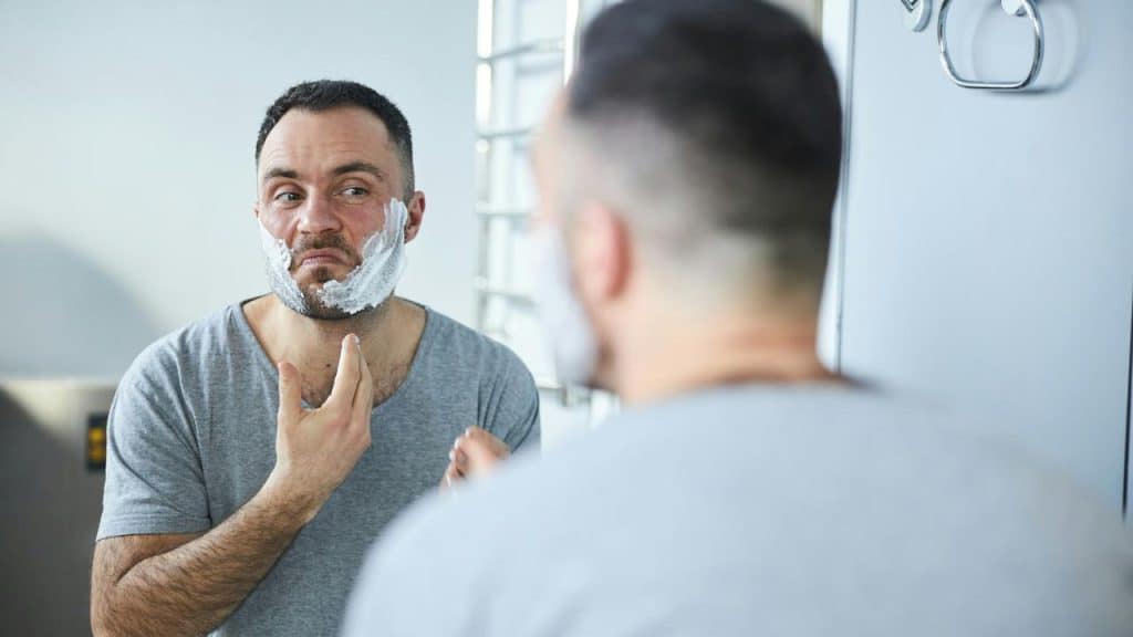 A man applying shaving cream to his face while looking in the mirror.