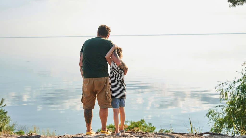 A father with his arm around his child looking out over a calm lake.
