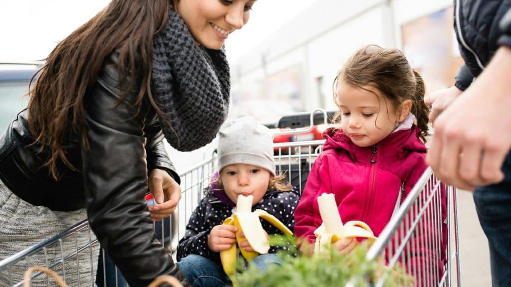 A woman shopping with two children eating bananas in a grocery cart.