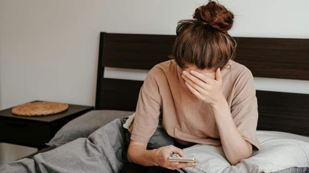 A woman sitting on a bed covering her face while holding a phone.