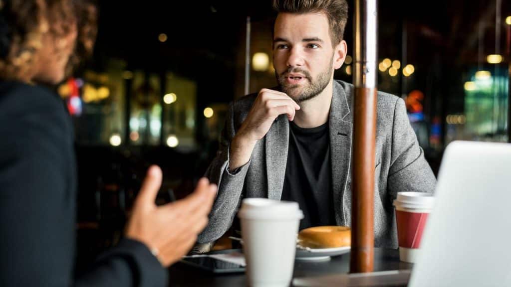 A man in a gray blazer talking at a café table.