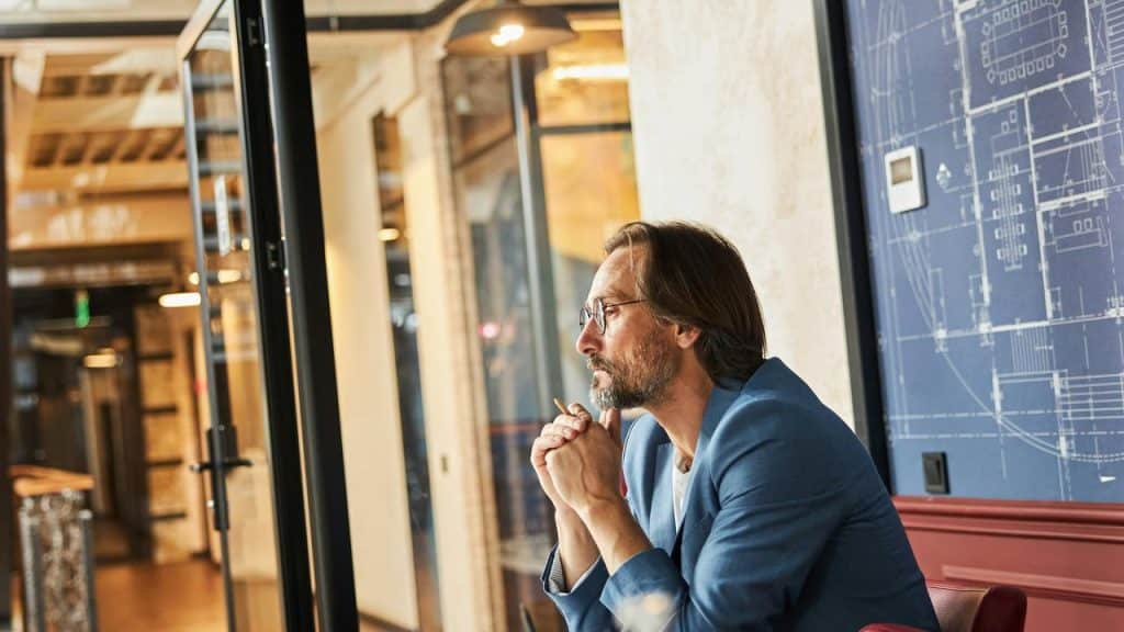 A man in a blue jacket sitting thoughtfully in an office.