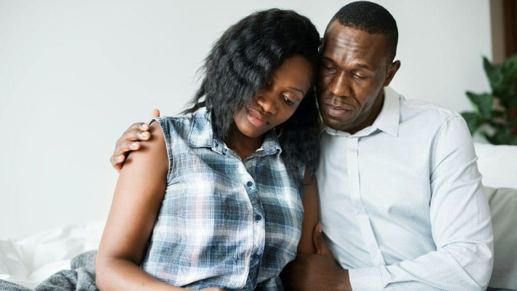 A sad couple is embracing each other on a bed, with the man's arm around the woman's shoulder.