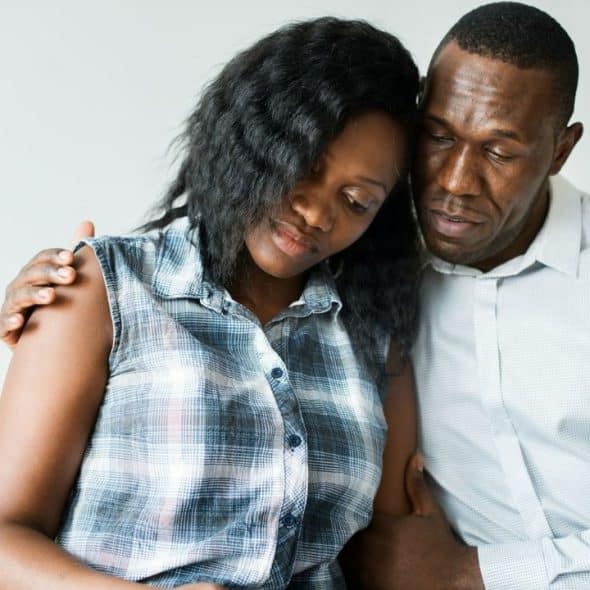 A sad couple is embracing each other on a bed, with the man's arm around the woman's shoulder.