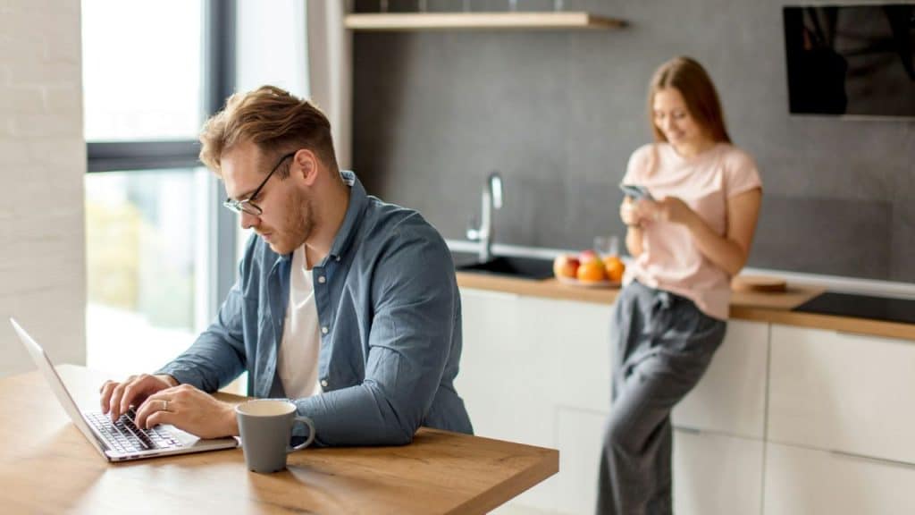 A man works on his laptop at a kitchen table while a woman looks at her phone in the background.