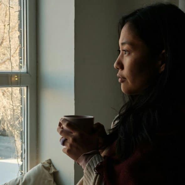 A woman with dark hair holds a mug and looks thoughtfully out a window.