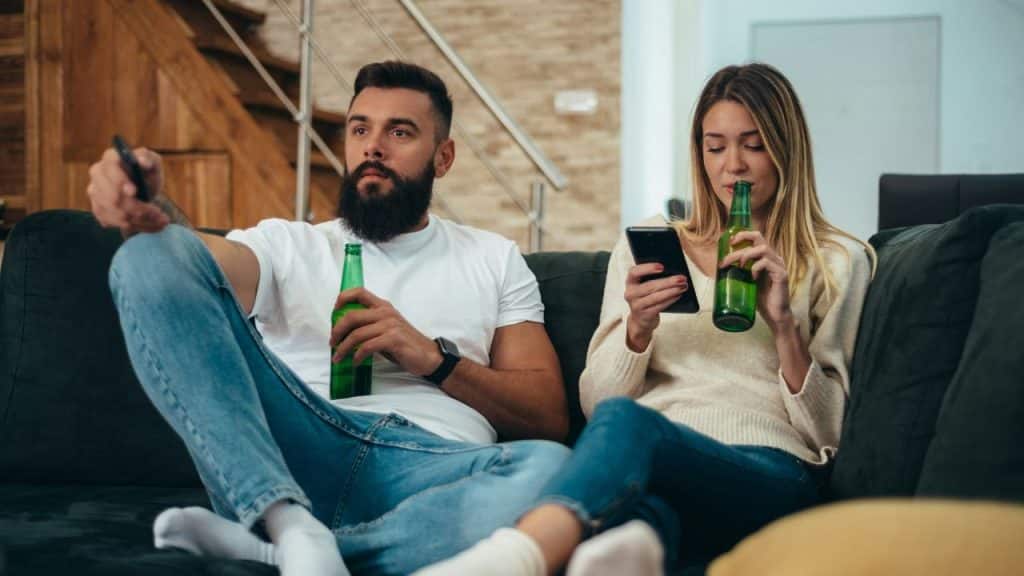 A couple sits on a couch, each holding a bottle of beer and looking at a different screen.