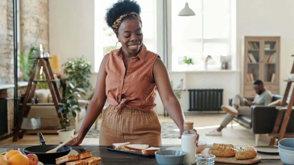 A smiling woman prepares food in the kitchen while a man is seen relaxing on a couch in the background.