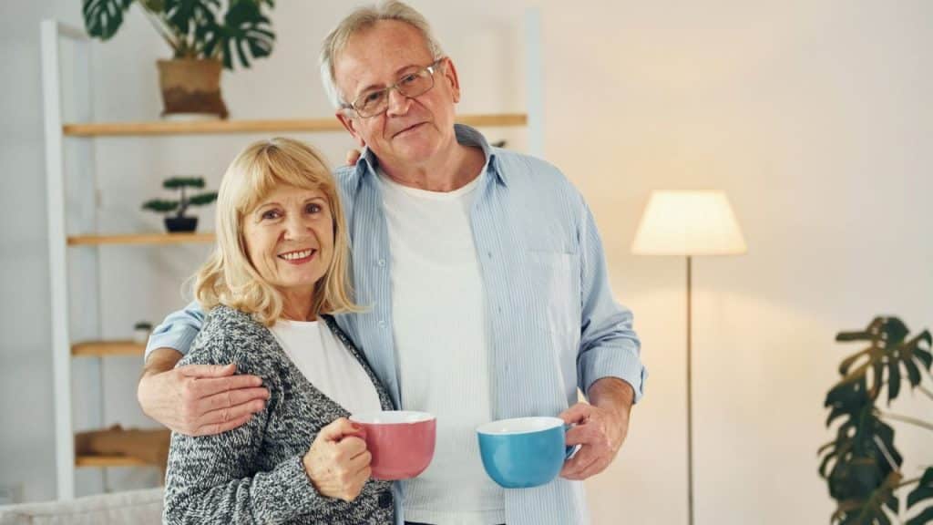 A smiling elderly couple holding mugs stands in their living room.
