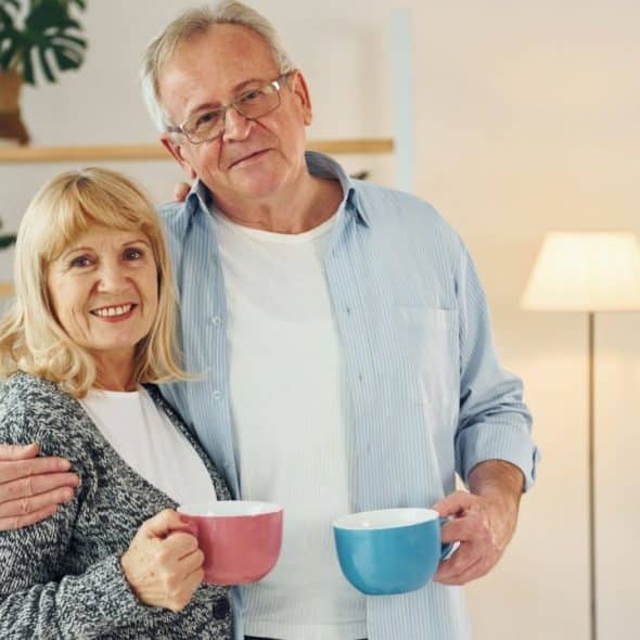 A smiling elderly couple holding mugs stands in their living room.