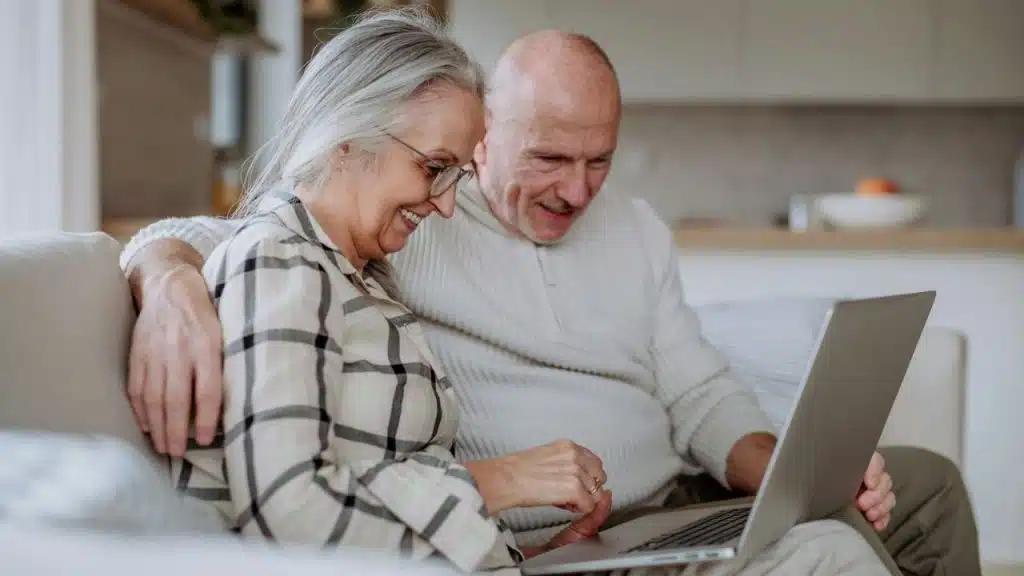 A happy elderly couple is sitting on a sofa and looking at a laptop together.