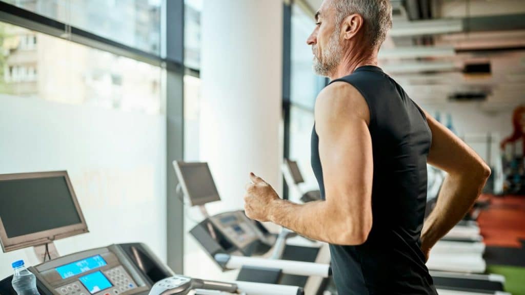 A gray-haired man in a black tank top is running on a treadmill at a gym.