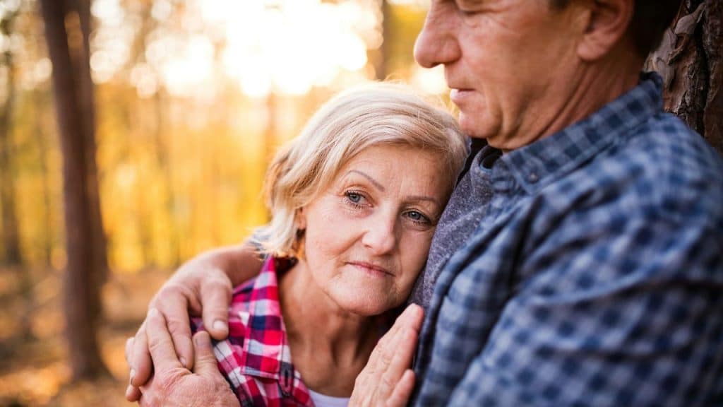 A senior couple embraces each other in a sunlit forest.