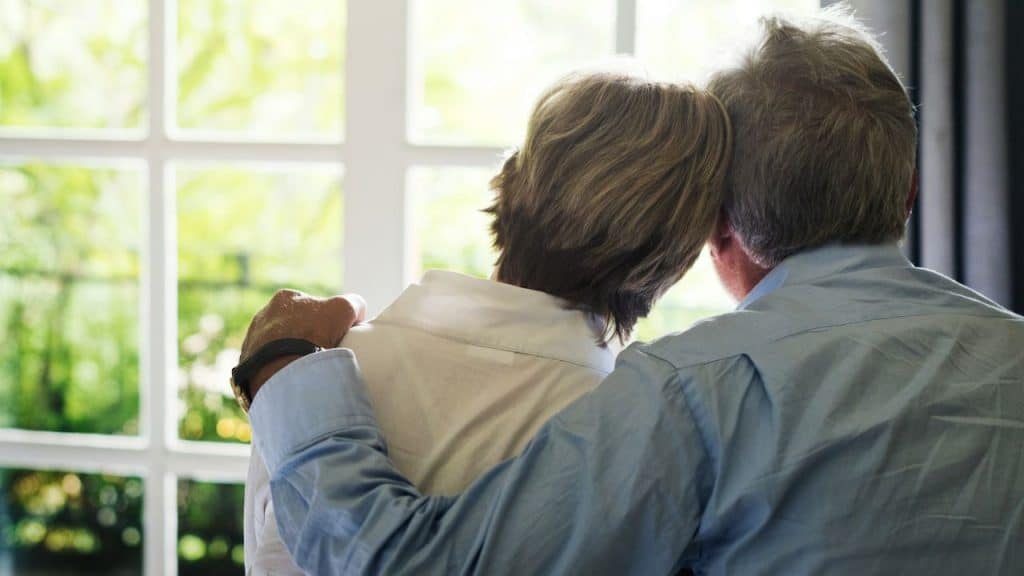 A loving elderly couple with their backs to the camera looks out a window.