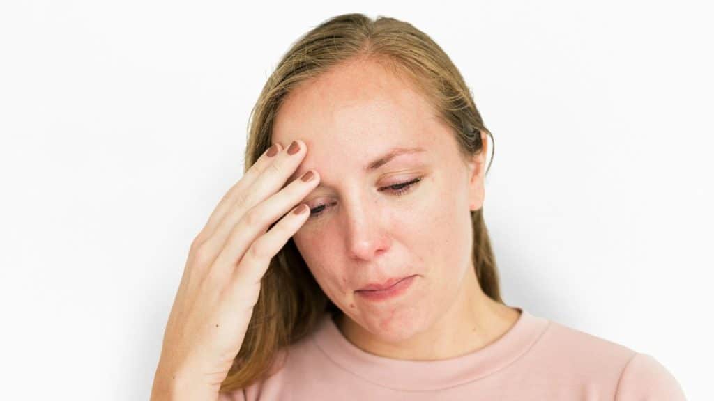 A sad-looking, long-haired woman in a light pink shirt holds her forehead and looks down.