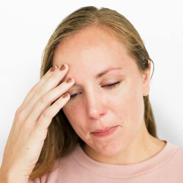 A sad-looking, long-haired woman in a light pink shirt holds her forehead and looks down.