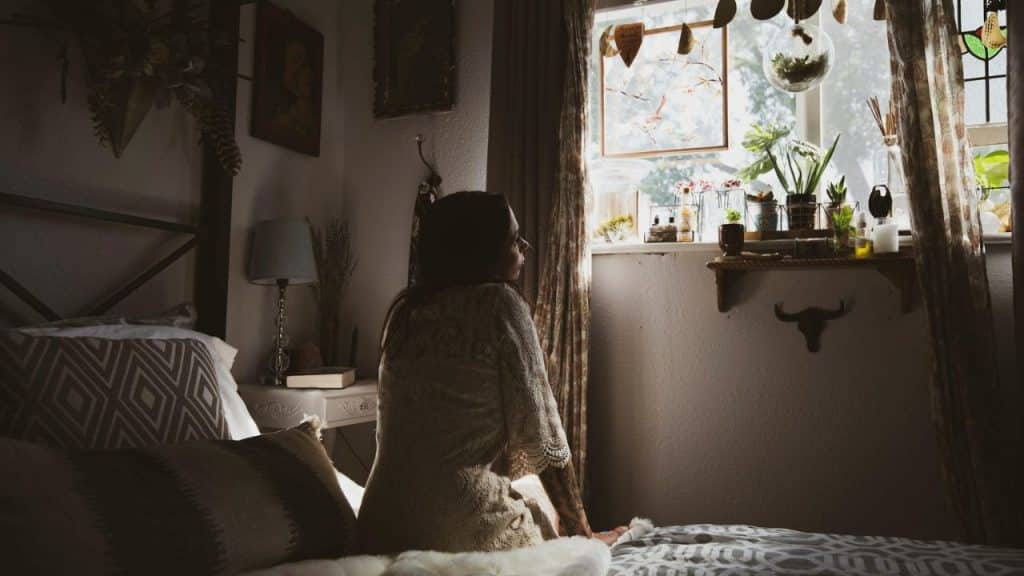 A serious woman in a white lace dress sits on a bed and looks out a sunlit window.