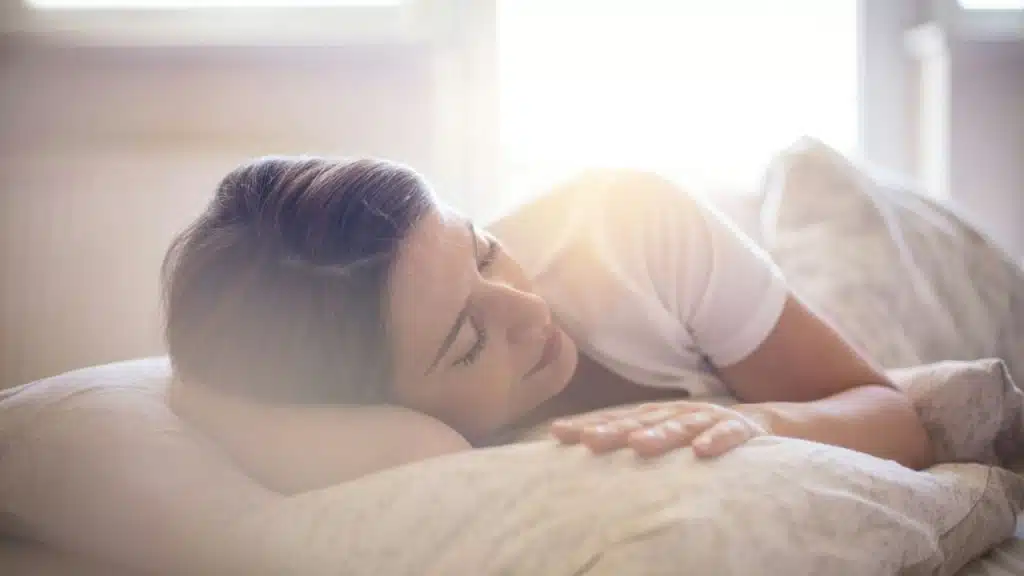A woman with her eyes closed is lying in bed, a hand on her pillow.
