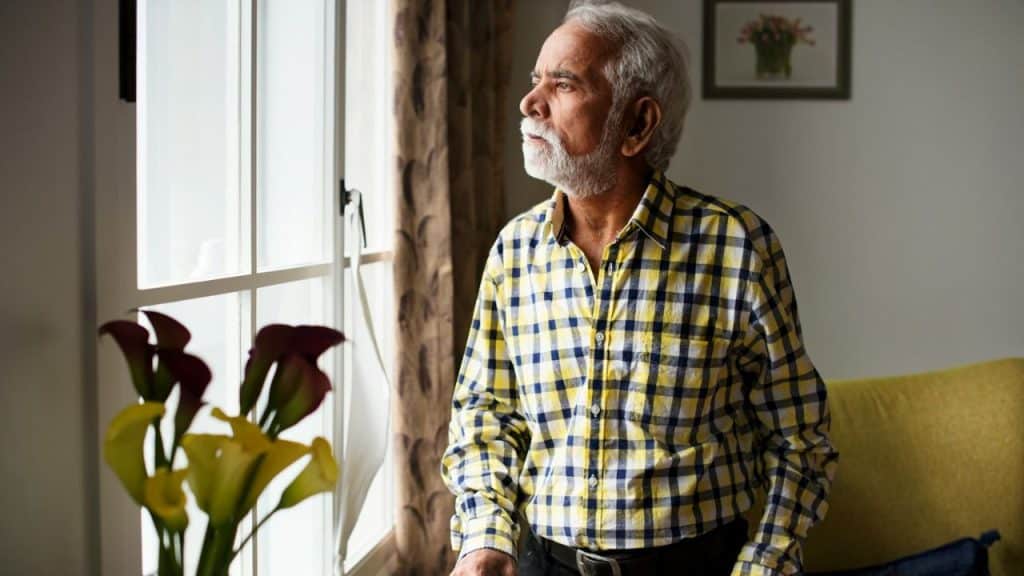 A thoughtful, gray-haired man in a yellow plaid shirt is looking out a window.