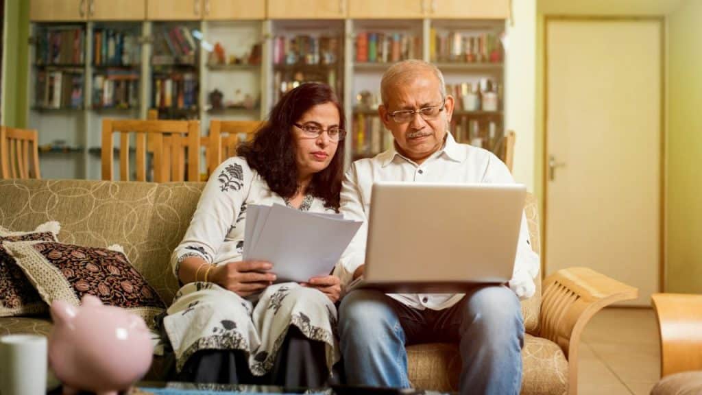 A serious-looking elderly couple is sitting on a sofa and looking at a laptop.