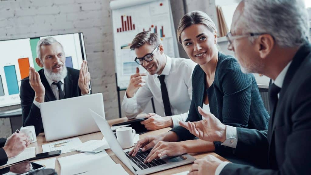 A business meeting is taking place, with a group of diverse professionals smiling and gesturing around a laptop.