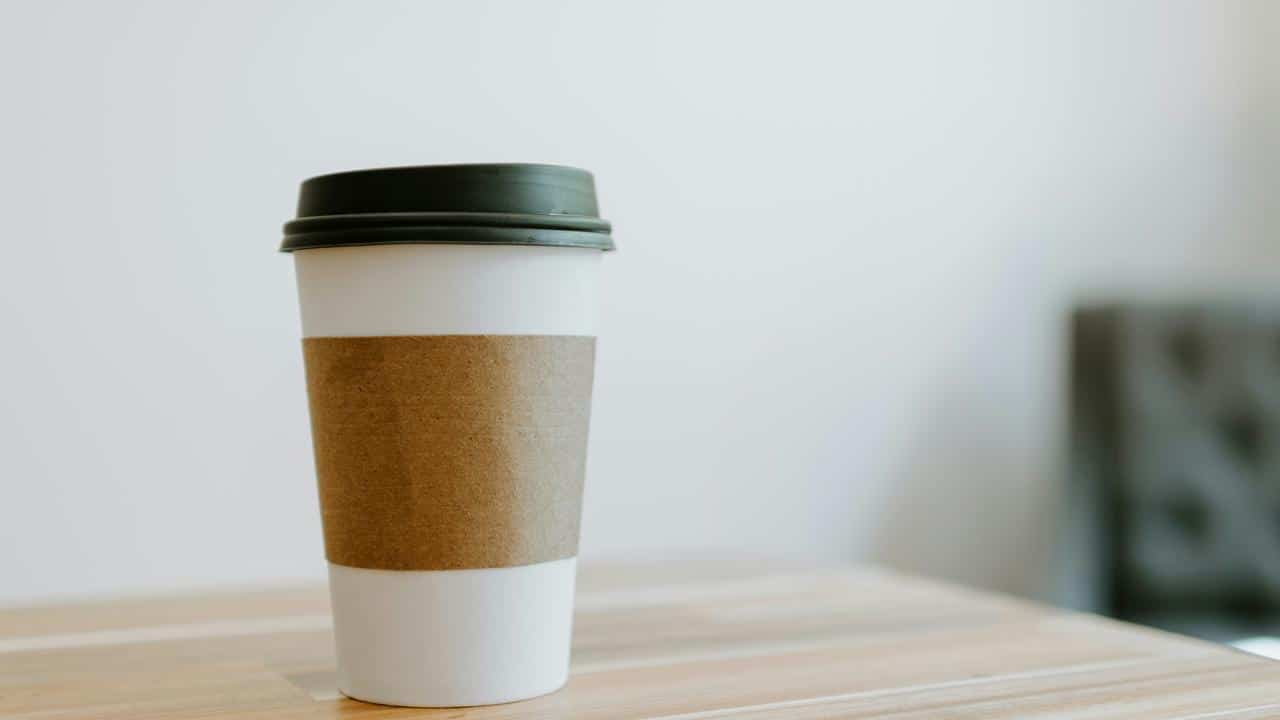 A disposable coffee cup with a black lid and a cardboard sleeve on a wooden table.