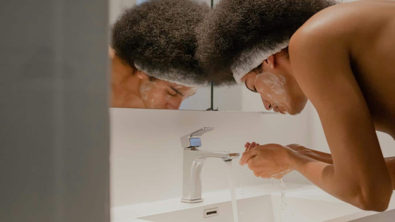 A person washing their face at a sink with a headband and facial cleanser.