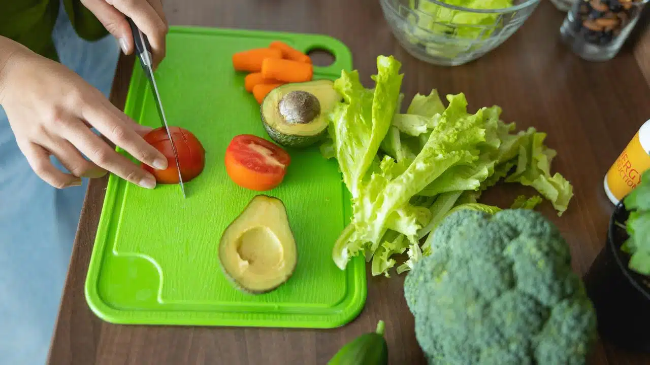 A person slicing a tomato on a green cutting board with assorted vegetables around.