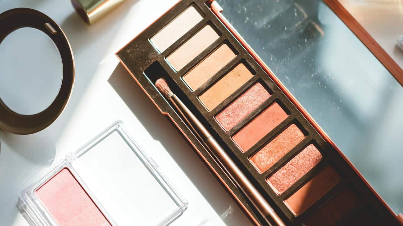 An eyeshadow palette with warm shades and a makeup brush next to other cosmetics.