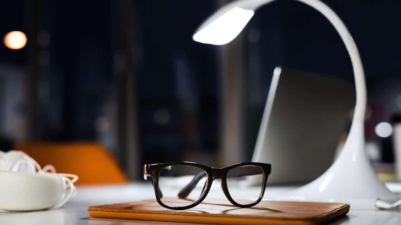 A pair of eyeglasses on a desk under a lamp.