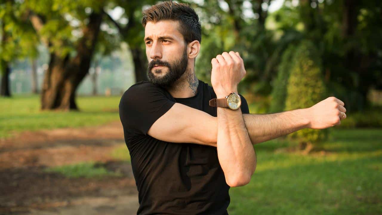 A man stretching his arm in a park during exercise.