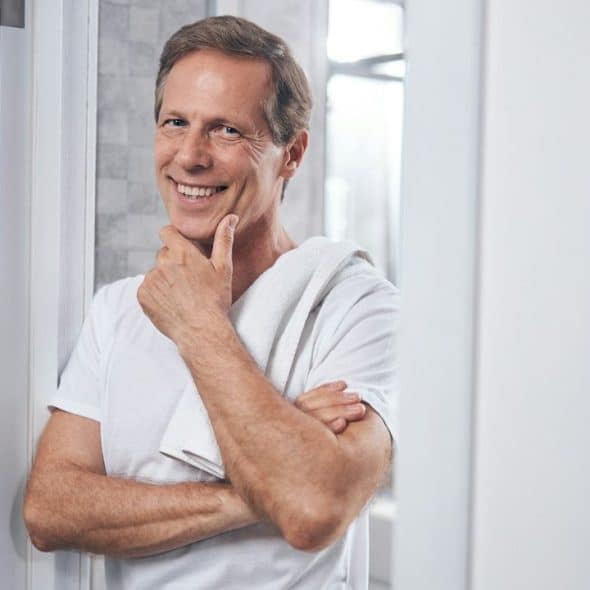 A happy, middle-aged man with a white towel over his shoulder is smiling and touching his chin.