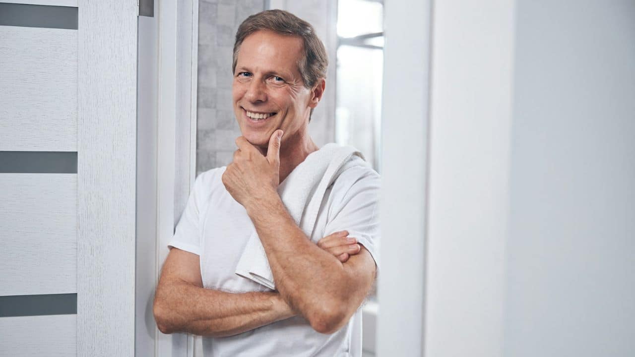 A happy, middle-aged man with a white towel over his shoulder is smiling and touching his chin.